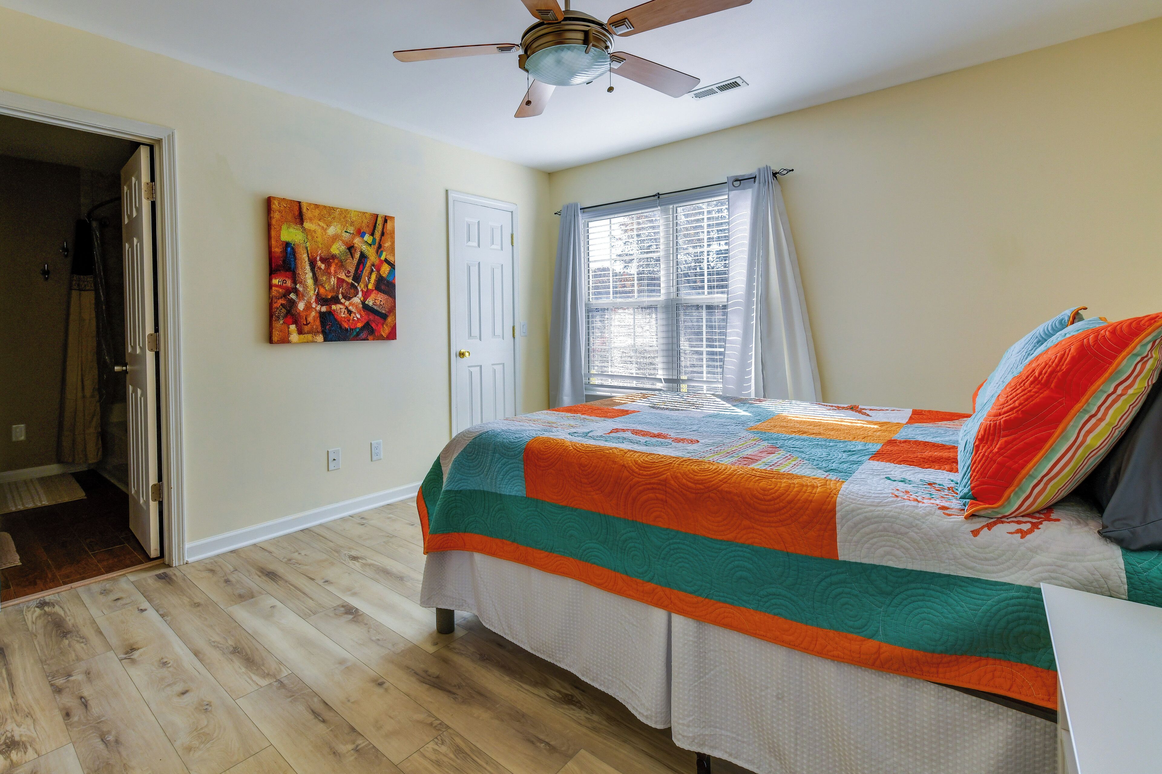 A bedroom with a bed, a ceiling fan, a window with curtains, and a door leading to another room.