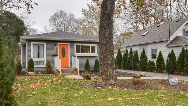 Une maison d’habitation à l’extérieur gris, avec une porte d’entrée orange et un petit porche agrémenté de plantes en pot.