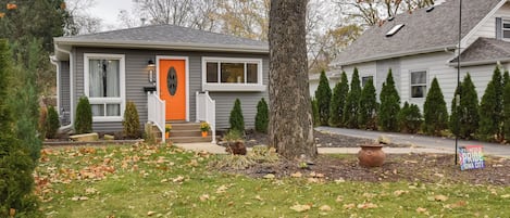 A residential house with a gray exterior, an orange front door, and a small porch with potted plants.