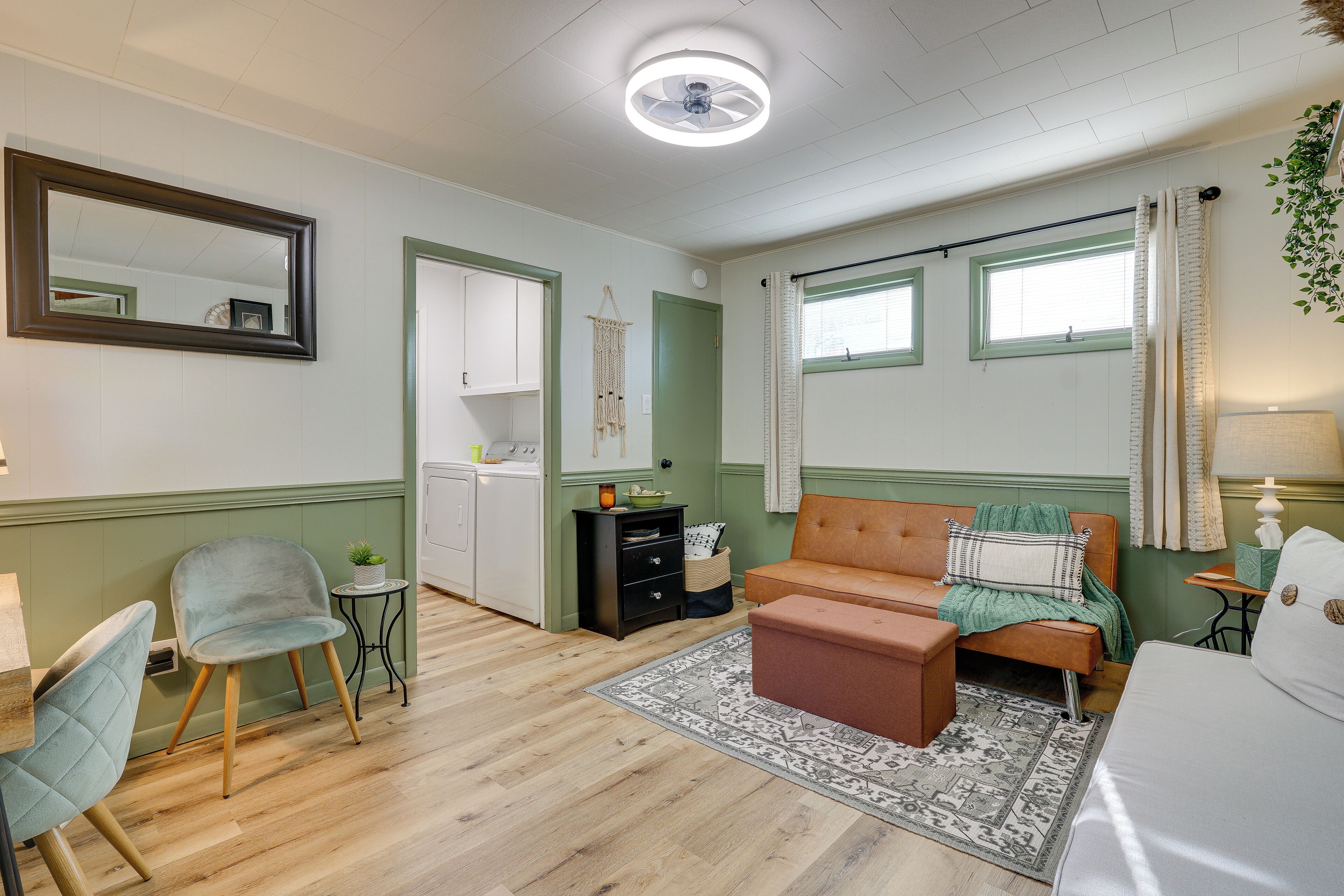 A room with a wooden floor, a sofa, a chair, a small table, a mirror, and a window with curtains.