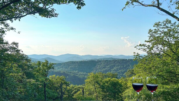 Una terraza de madera con dos copas de vino tinto, con vistas a un paisaje montañoso.