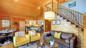 Interior acogedor de una cabaña con escalera de madera, comedor y sala de estar amoblada con sillas amarillas y un sofá marrón.