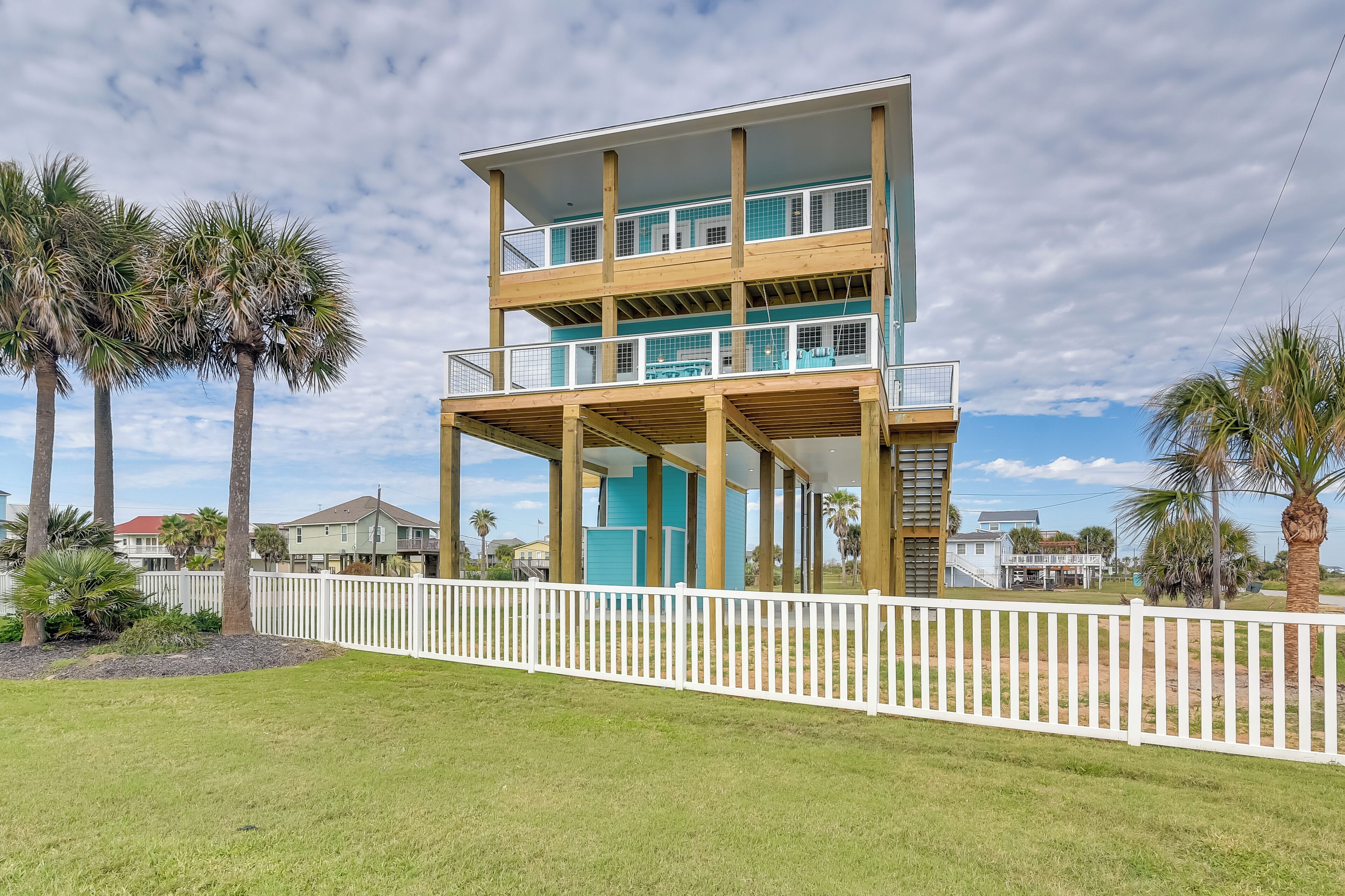 A multi-story building with balconies, surrounded by a white picket fence, palm trees, and a grassy area.