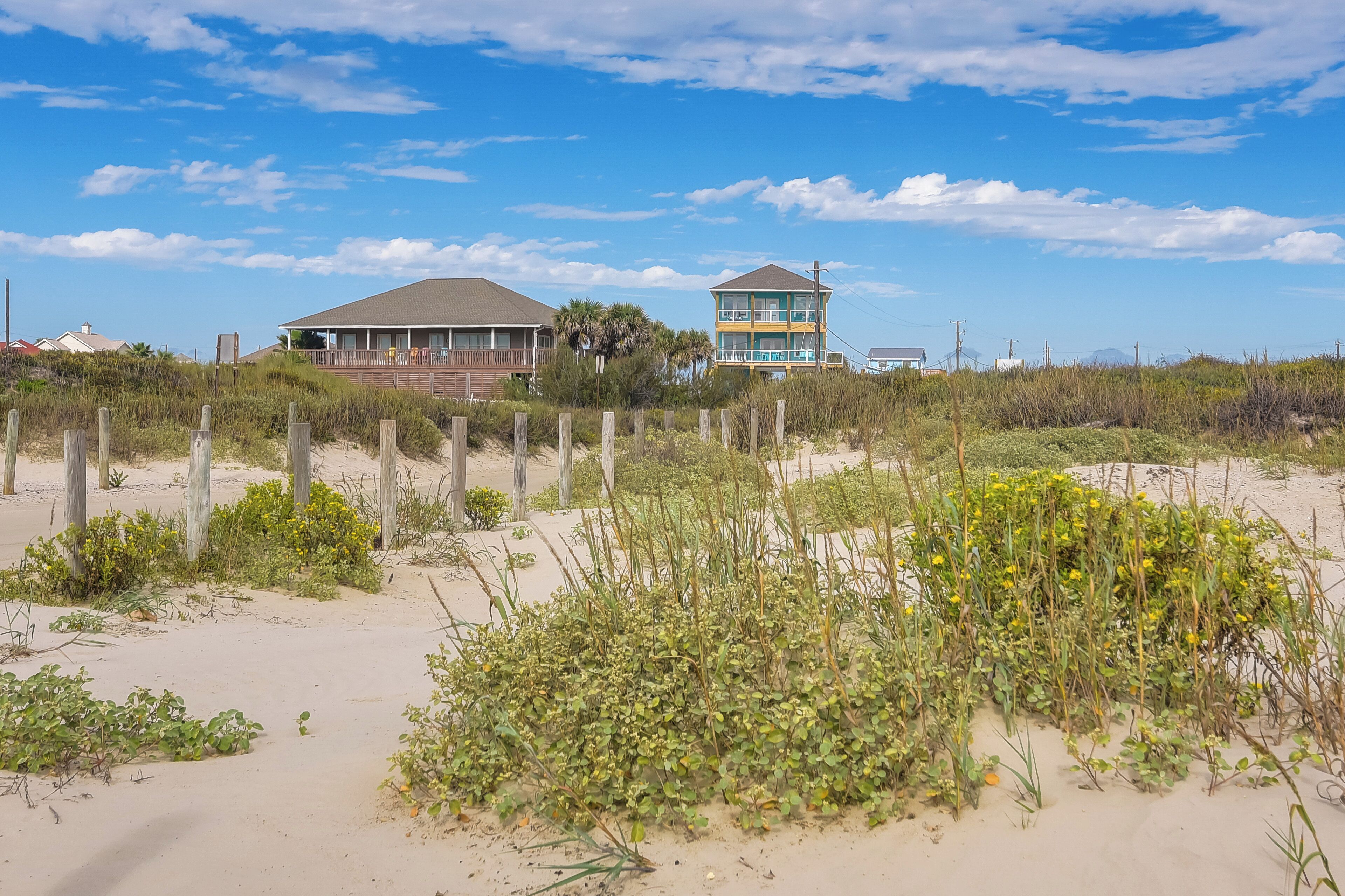 A beach house with a fenced yard and a sandy beach in the background.