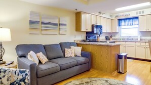 Ein modernes Wohnzimmer mit einem grauen Sofa, einem gemusterten Sessel und einer Küchenzeile mit Holzausziehtüren und einem Edelstahl-Mülleimer.