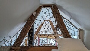 Une petite chambre mansardée de forme triangulaire, avec un lit en bois, un hamac et une fenêtre protégée par un moustiquaire.