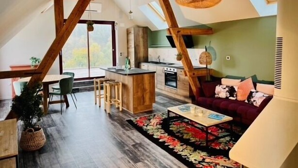 Un loft spacieux avec de hauts plafonds, des poutres apparentes, un coin cuisine et un espace salon confortable agrémenté d’un tapis à motifs floraux.