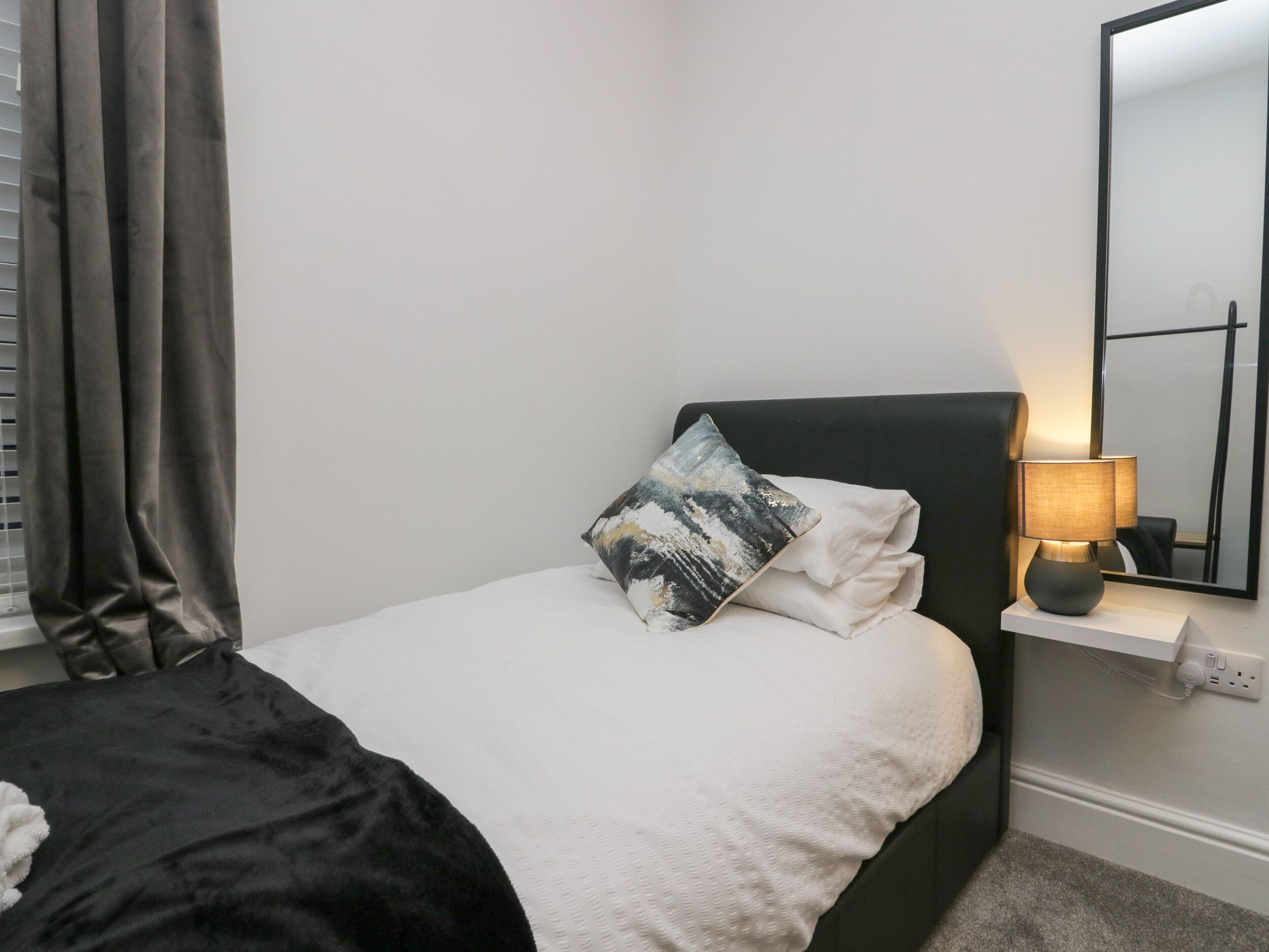 A bedroom with a bed, a bedside lamp, a mirror, and a window with curtains.