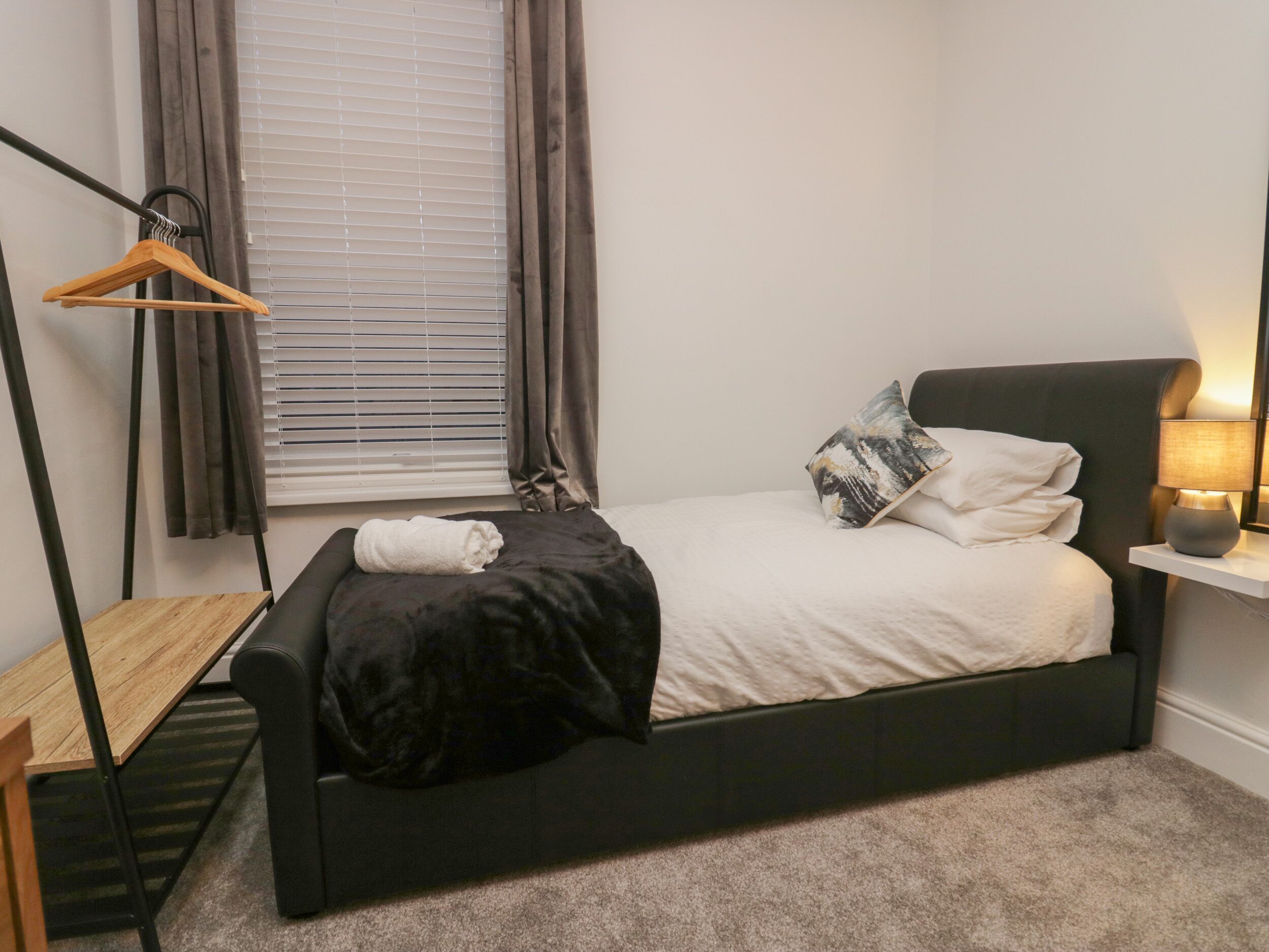 Un dormitorio con una cama negra, una mesita de noche, una lámpara, un perchero de madera colgante y una ventana con persianas.