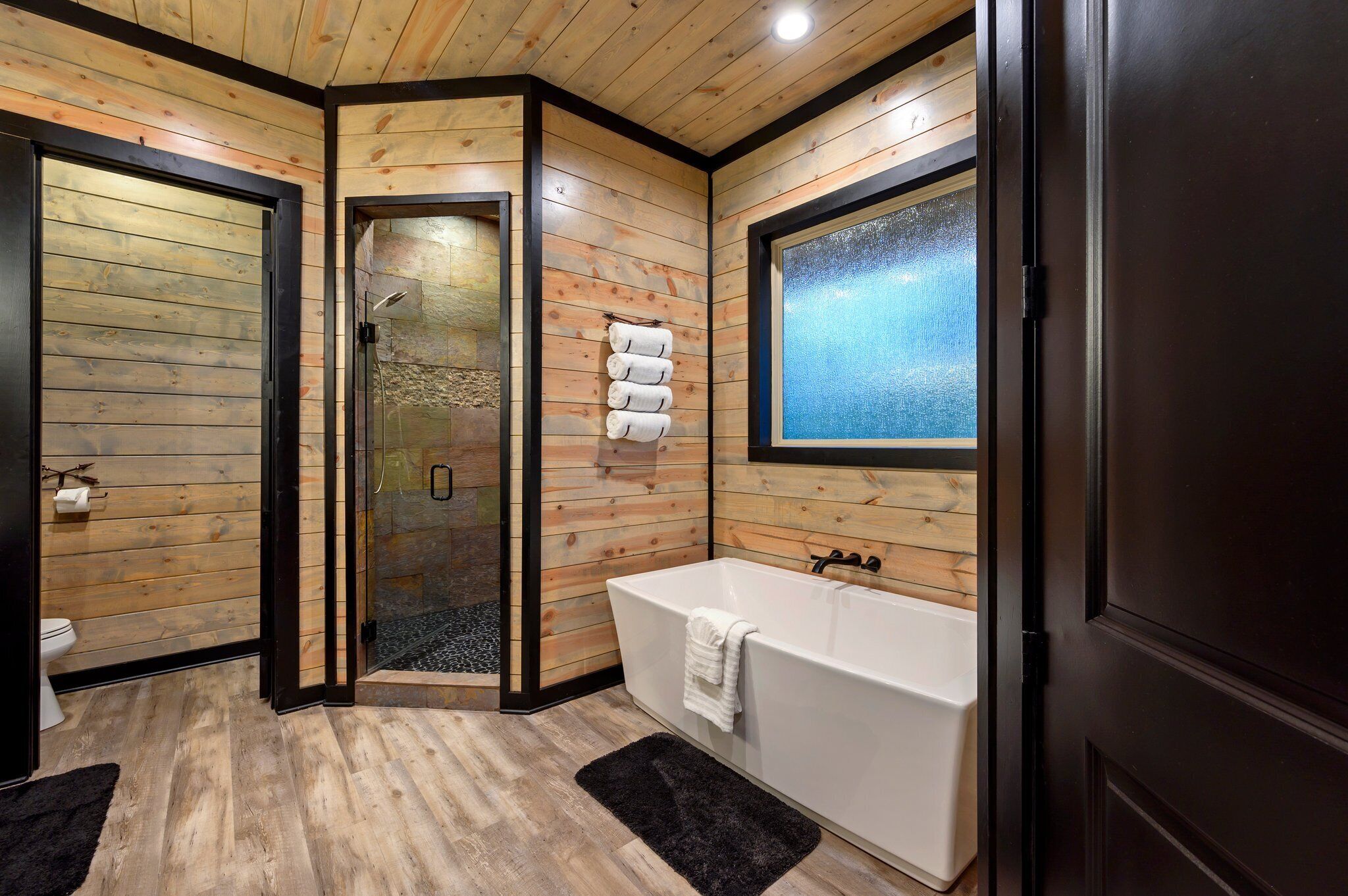 A modern bathroom with wooden walls, a glass shower enclosure, a bathtub, and a large window.