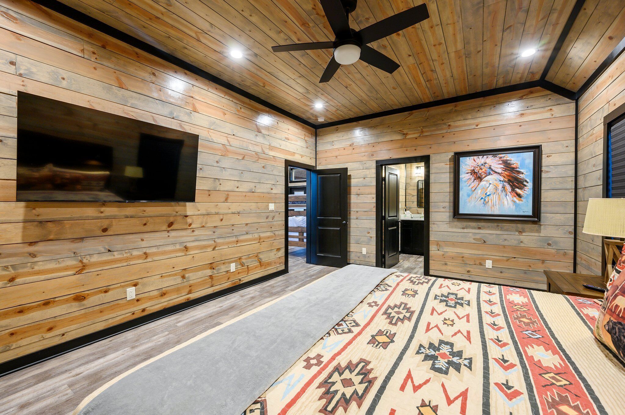 A room with wooden panel walls, a ceiling fan, a large flat-screen TV, a patterned rug, and a painting on the wall.