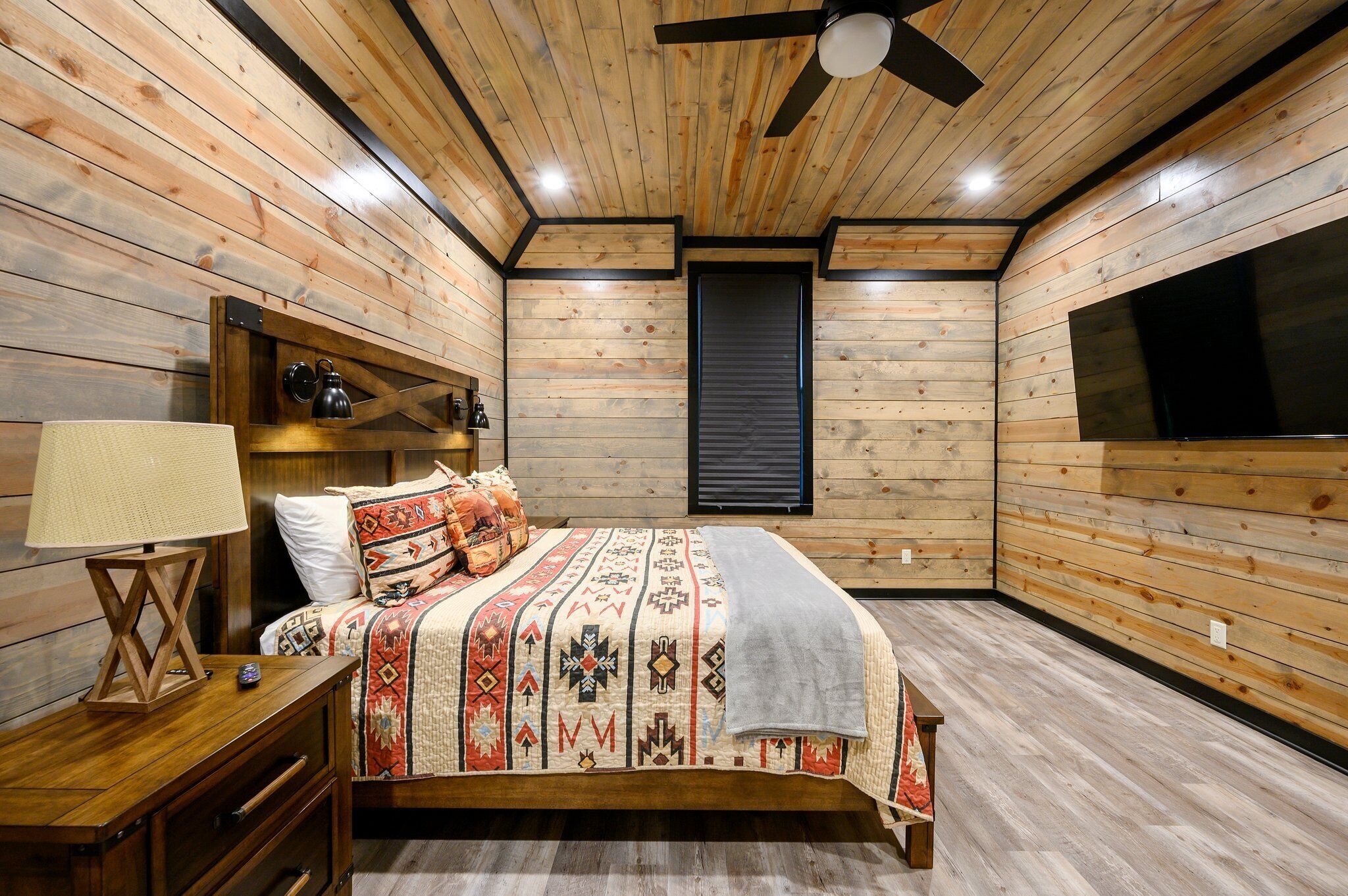 A wooden-paneled bedroom with a bed, nightstand, lamp, and a ceiling fan.