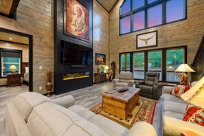 A spacious living room with a fireplace, comfortable seating, and large windows offering views of the outdoors. - Guests Favorite + Pool Table  + 2 Decks + 4 King Bedrooms + Bunk Loft + XL deck (Broken Bow)