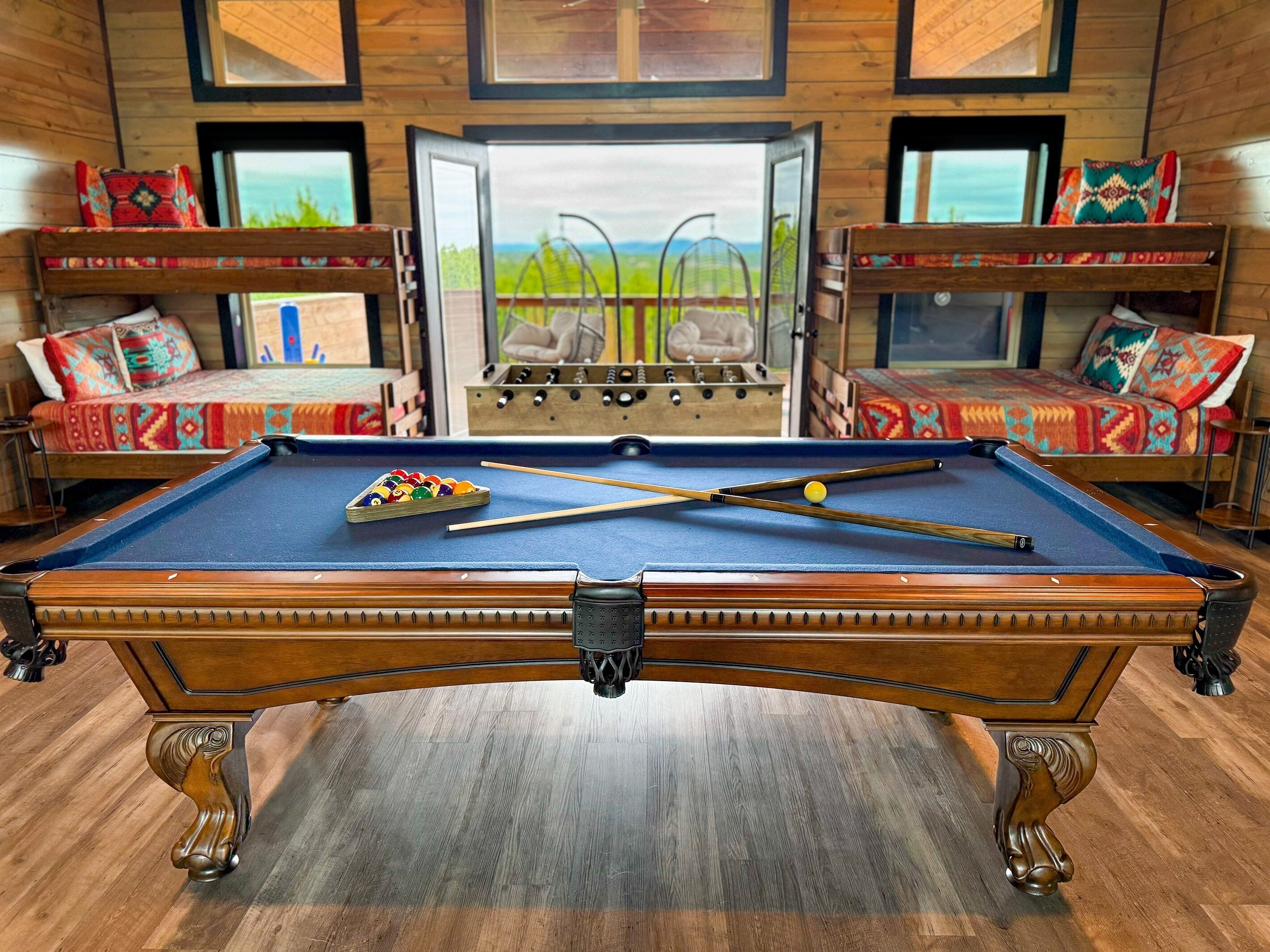 A pool table in a room with wooden walls and a view of the outdoors. There are two sofas with colorful cushions and a foosball table in the background.