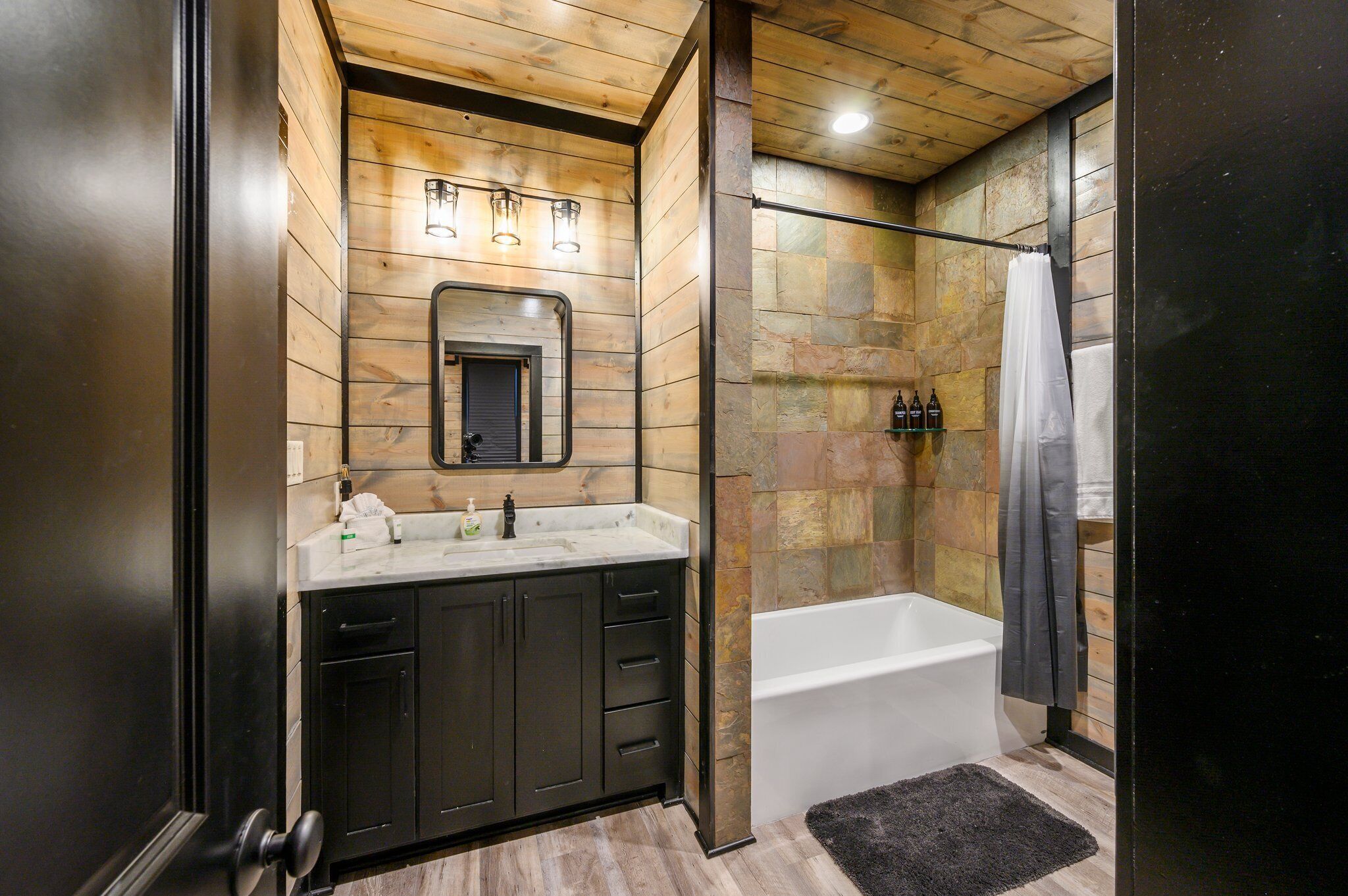 A modern bathroom with a wooden ceiling, a large mirror, a bathtub, and a stone wall.