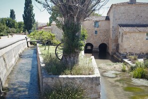 Un étroit canal où l’eau s’écoule, bordé d’un arbre et d’une roue, et entouré de bâtiments en pierre dotés d’entrées en arc de cercle.