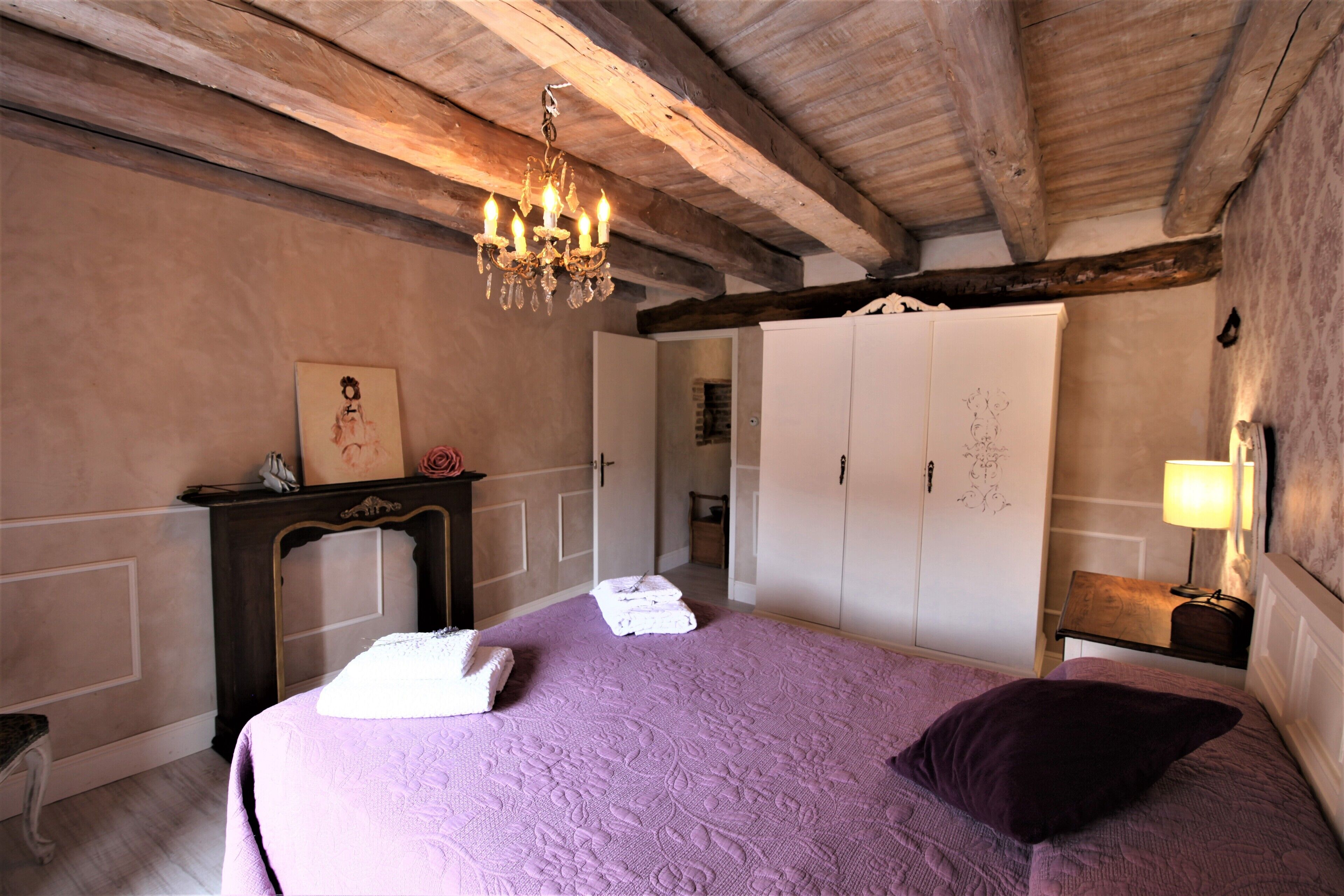 Quarto com teto de madeira, um candelabro, uma cama com colcha roxa, um guarda-roupa branco e uma mesa de cabeceira com abajur.