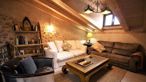 A cozy living room with a white sofa, a brown sofa, a wooden coffee table with chess pieces, a lamp, and a bookshelf against a stone wall.