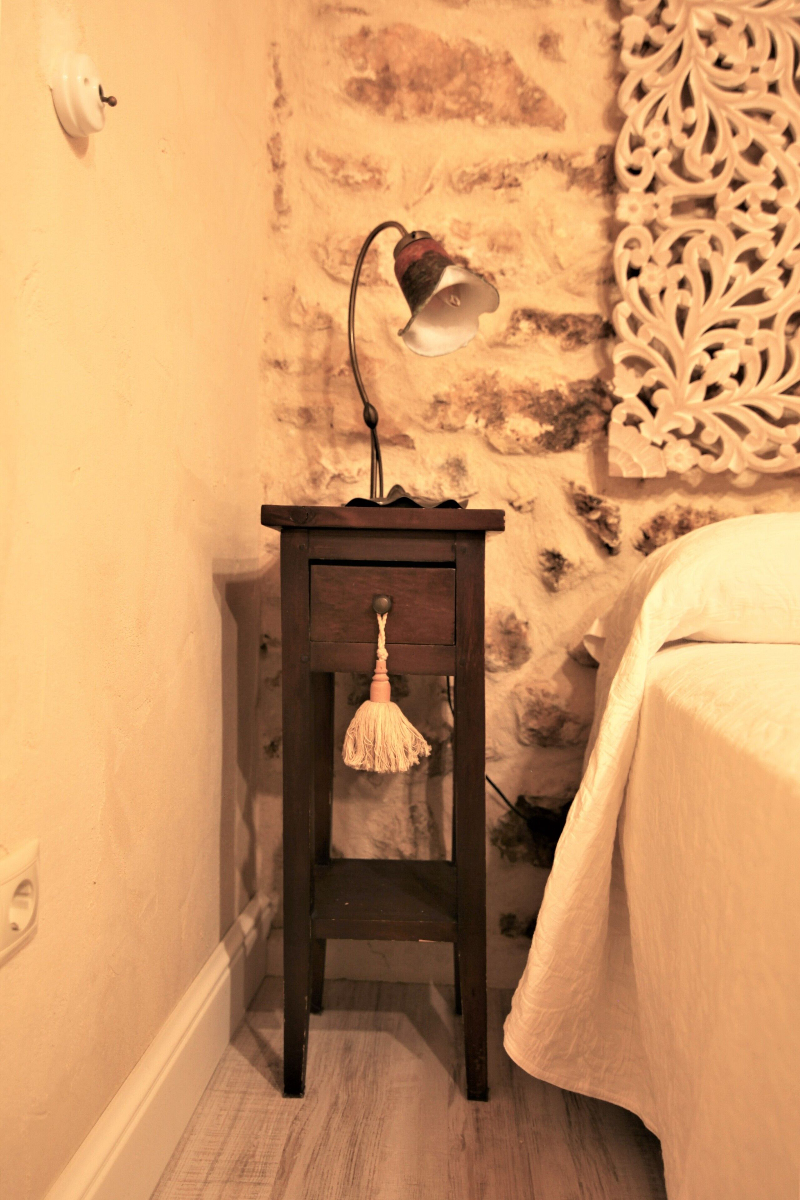 Una pequeña mesita de noche con una lámpara, una pared decorativa y una cama con colcha blanca.