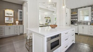 Une cuisine moderne avec des meubles blancs, un îlot central doté d’un four encastré, un éclairage suspendu et une vue sur la salle à manger.