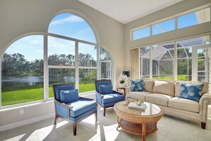 A spacious room with large windows offering a view of a lake and greenery. There are two blue armchairs and a beige sofa with decorative pillows. A round wicker coffee table is in the center.