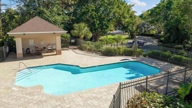 Une piscine avec une terrasse carrelée, un espace salon couvert et une cour clôturée agrémentée d’arbres, avec une voiture visible au loin.