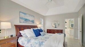 A bedroom with a bed, bedside tables, a nightstand, a ceiling fan, a painting, a mirror, and a door leading to another room.