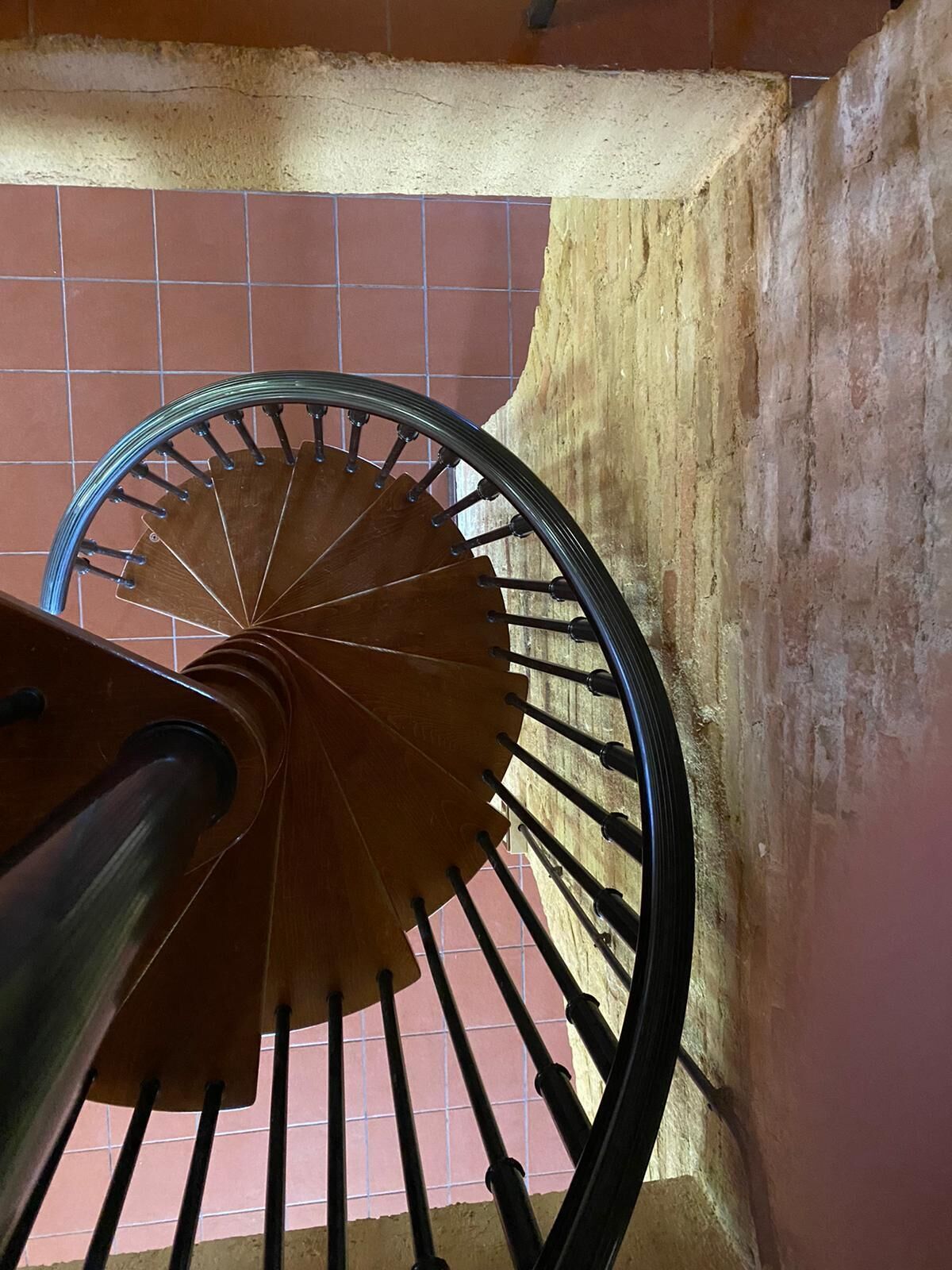 En spiraltrappa med ett träräcke och svarta spjälor, som leder ner till ett klinkergolv.