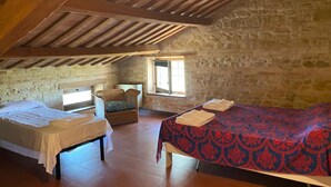 A rustic attic room with two beds, a wicker chair, and a stone wall. - Attic Room with Private Bathroom in a Historic Villa Farmstay (Marche)