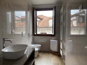 A modern bathroom with a large oval sink, a toilet, and a bidet. There is a window providing a view of buildings with tiled roofs. - Ca' Magna Apartment  (Venezia)