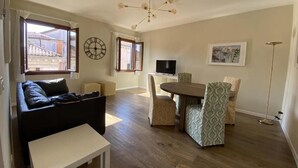 A living room with a sofa, a small table, a dining table with chairs, a TV, a clock, and a floor lamp. - Ca' Magna Apartment  (Venezia)