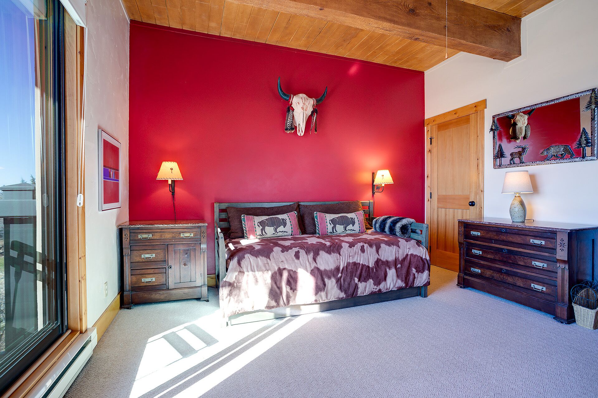 A bedroom with a red wall, a bed with patterned bedding, two bedside lamps, a wooden dresser, and a door with a mirror.