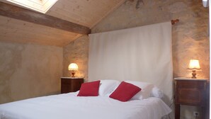 A bedroom with a wooden ceiling, a bed with white bedding and red pillows, two bedside lamps, and a stone wall. - Cottage for 4 in a small village near Uzès, with garden, pool and airco (AUBUSSARGUES)