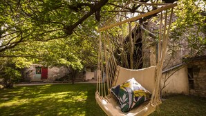 A hammock swing with a cushion, set in a lush garden with a stone wall and a building in the background. - Cottage for 4 in a small village near Uzès, with garden, pool and airco (AUBUSSARGUES)