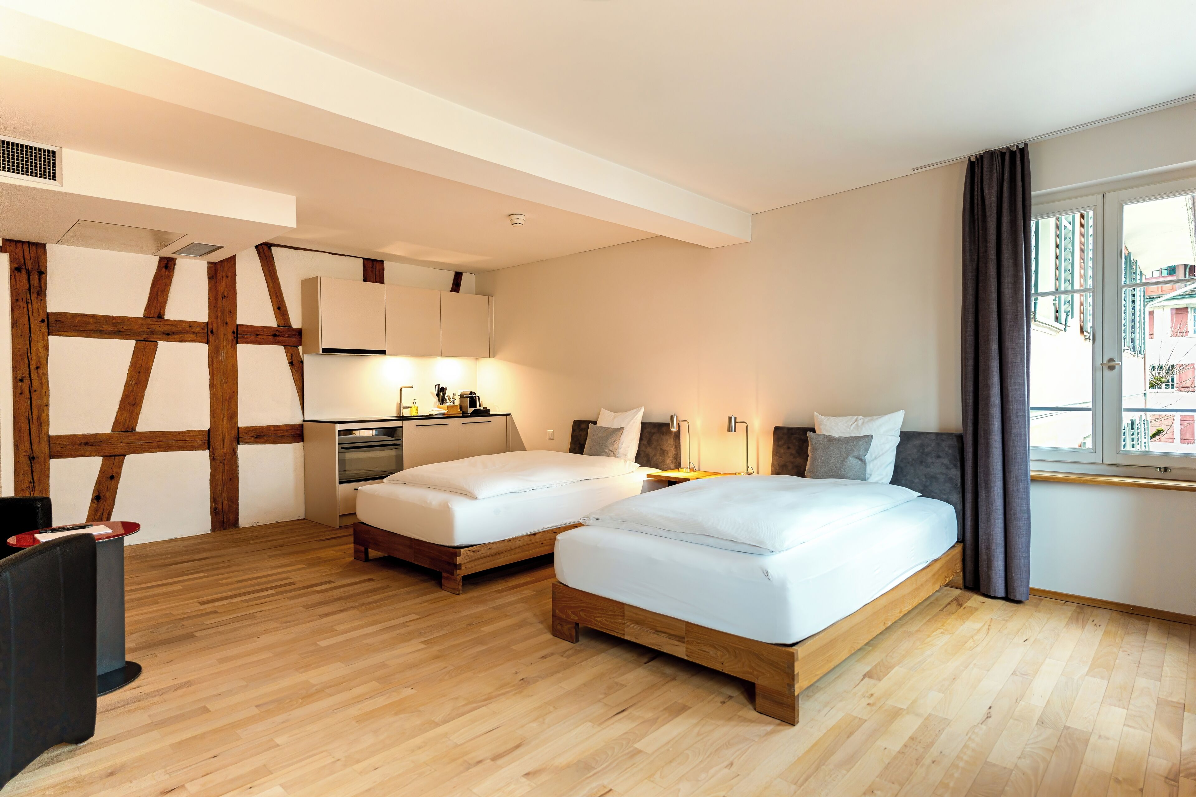 A modern hotel room with two beds, wooden flooring, a kitchenette, and a large window with curtains.