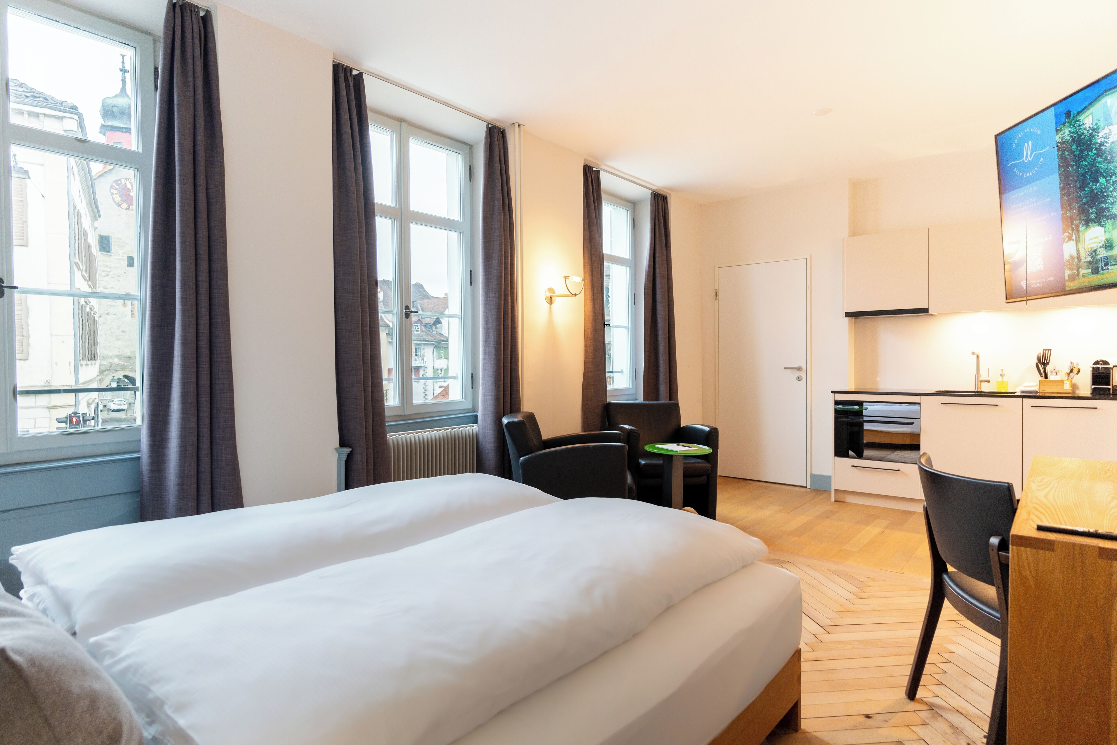 Une chambre d’hôtel avec deux lits, un coin repas comprenant une table et des chaises, un coin cuisine et une grande fenêtre avec des rideaux.