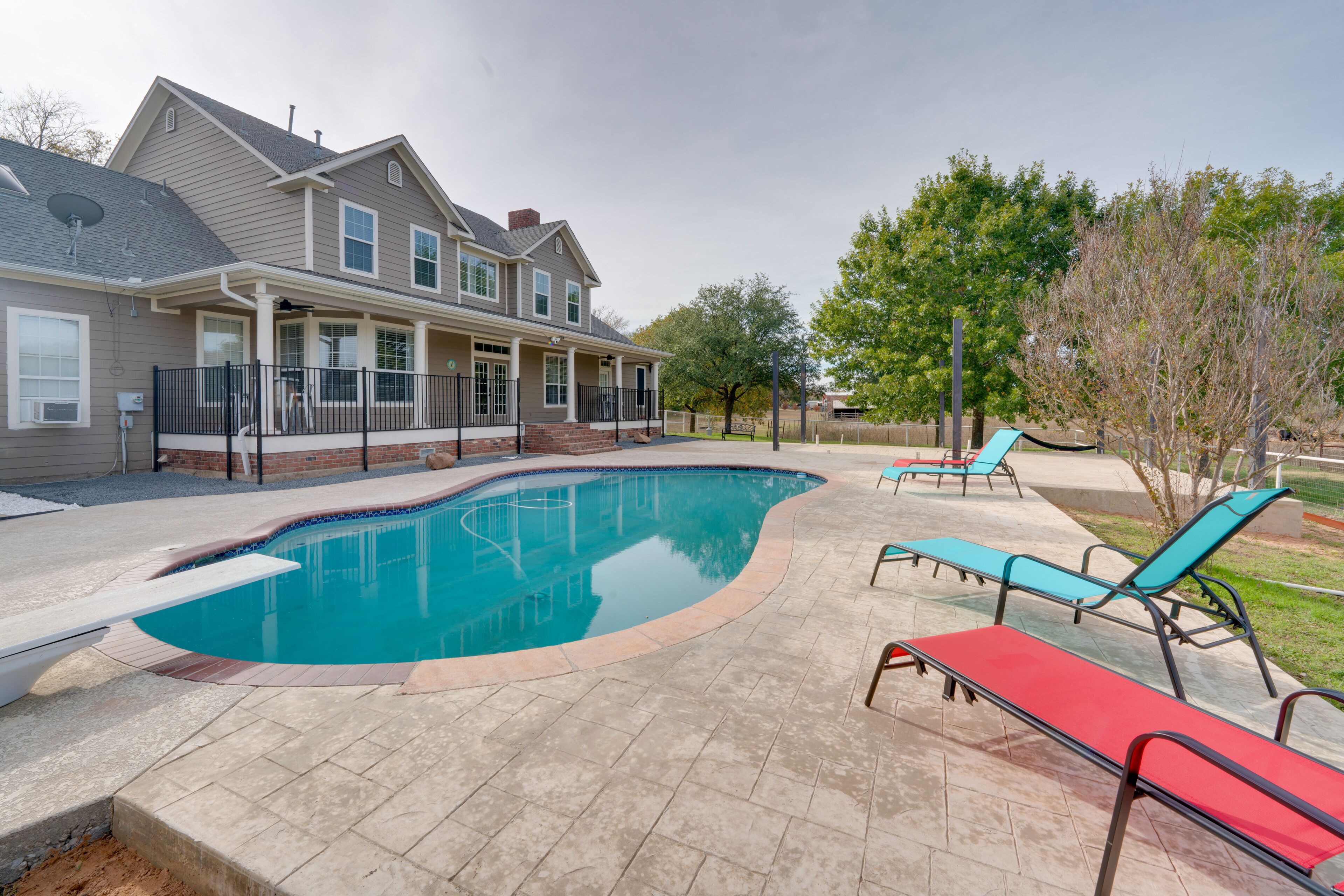 A residential swimming pool with a house in the background, surrounded by a patio area with lounge chairs.