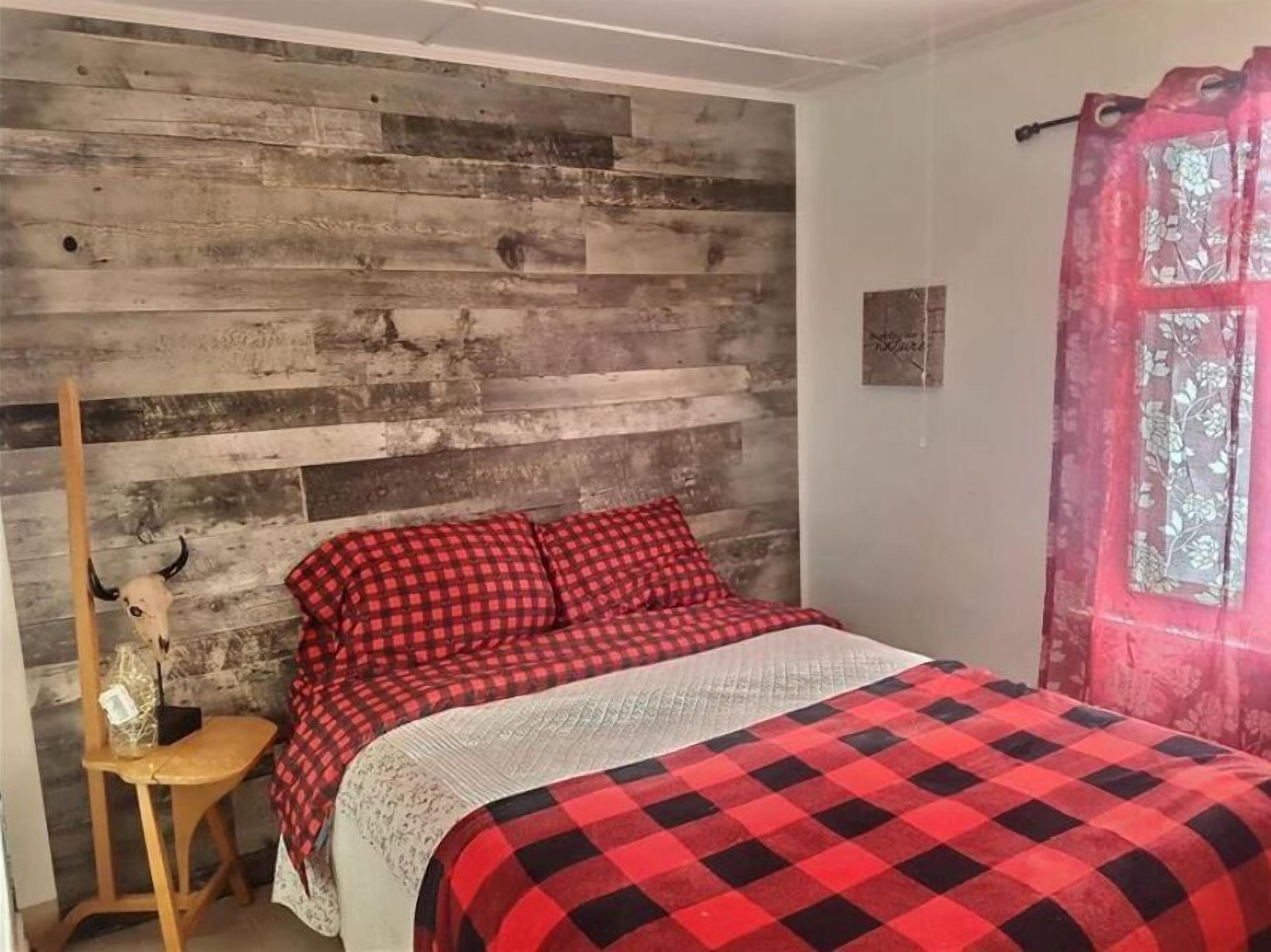 A bedroom with a wooden wall, a bed with red and black checkered bedding, a wooden chair, and a pink curtain.
