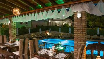 Un área de comedor al aire libre con mesas y sillas, una piscina y un espacio de estar cubierto con vista a la piscina.