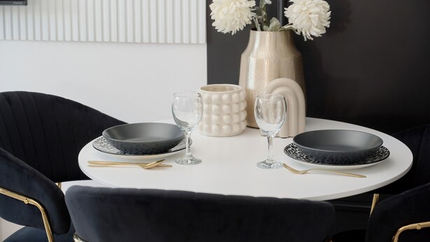 A dining table with black plates, clear glasses, and gold cutlery, set against a white wall with a decorative vase of white flowers. - Tastefully Arranged Studio by Renters (Wroclaw)