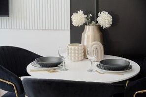 A dining table with black plates, clear glasses, and gold cutlery, set against a white wall with a decorative vase of white flowers. - Tastefully Arranged Studio by Renters (Wroclaw)