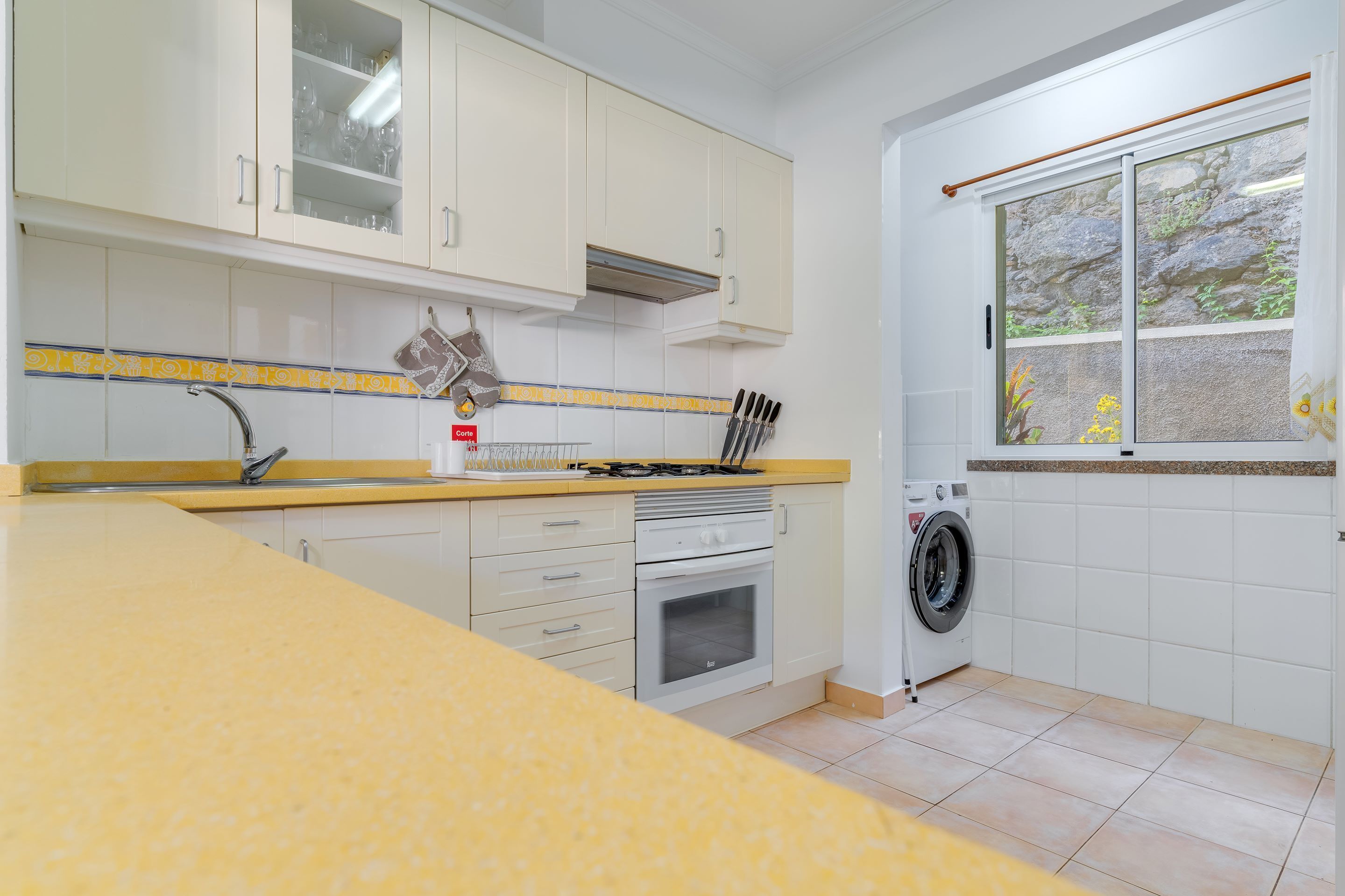Une cuisine avec des meubles blancs, un plan de travail jaune, une cuisinière, un évier et un lave-linge.