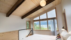 A bedroom with a large window, wooden ceiling beams, a bed, and a bedside table.