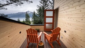 A wooden deck with two Adirondack chairs and a table, overlooking a lake and mountains.