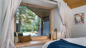 Makuuhuoneessa on suuri ikkuna, sinisellä peitolla varustettu sänky, puinen penkki ja seinällä oleva taulu.