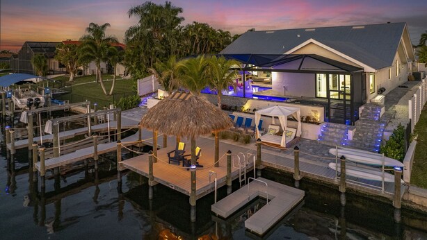 A waterfront property with a pool, dock, and thatched-roof seating area. - Kayaks, Boat Dock, Heated Pool & Spa, Outside Kitchen, Tiki Hut - Villa Pedro - Roelens Vacations (Cape Coral)