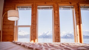 A room with a bed, a lamp, and three windows offering a view of snow-capped mountains and a lake. - Rustic Roots Seaside Blush Cabin (ADA) (Seward)