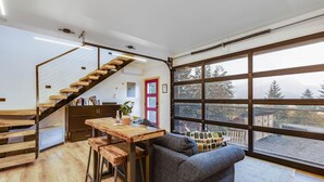 A modern kitchen with a wooden staircase, a dining area with stools, and a large window offering a view of the outdoors. - Salted Roots Purple Door Cottage with Ocean Views (Seward)