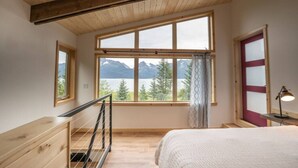 A bedroom with a large window offering a mountain view, a bed, a desk, and a chair. - Salted Roots Purple Door Cottage with Ocean Views (Seward)