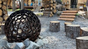 A rustic cabin with a unique spherical metal sculpture in the foreground, surrounded by wooden logs and tree stumps. - Salted Roots Purple Door Cottage with Ocean Views (Seward)
